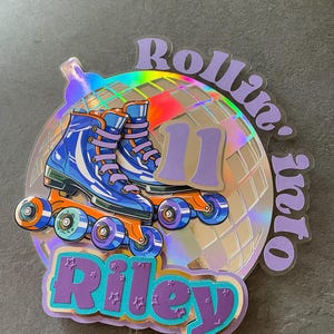 May include: A purple and blue cake topper with a disco ball, roller skates, and the text "Rollin' into Riley".