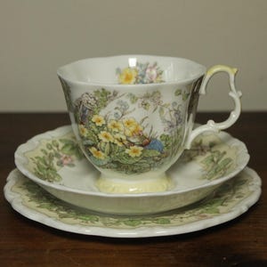 Puede incluir: Un juego de té vintage que consta de una taza, un platillo y un plato lateral. Las piezas de porcelana blanca presentan un diseño floral y paisajístico con acentos amarillos, verdes y azules. La taza tiene un asa curva.