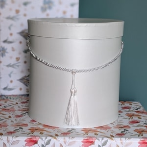 May include: A white, cylindrical gift box with a silver rope handle and tassel. The box sits on a floral patterned surface with a matching box in the background. The background is a solid teal color.