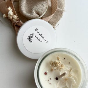 Peut inclure: Une bougie blanche dans un pot en verre transparent, décorée de fleurs séchées et de paillettes. La bougie est à côté d'un couvercle blanc avec le texte "Nous allons nous marier!" et un motif floral. Un ruban et du tissu sont en arrière-plan.