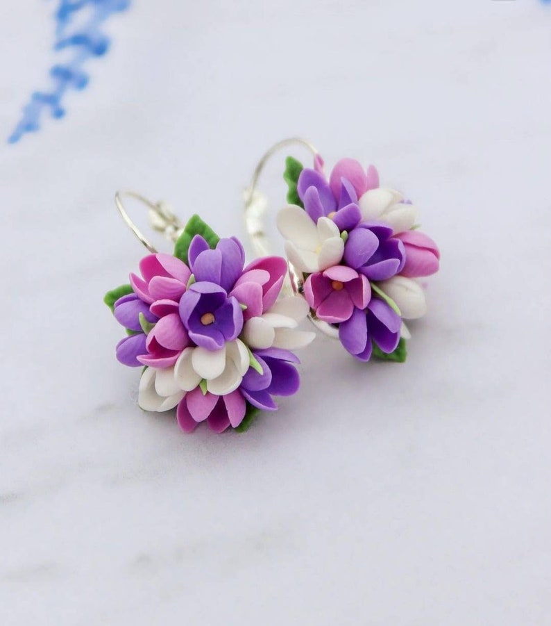 May include: A pair of silver hoop earrings with clusters of purple, pink, and white polymer clay flowers. The flowers are arranged in a bouquet-like shape.