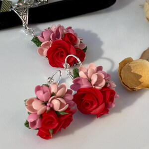 Peut inclure: Un ensemble de bijoux compos&eacute; d'un tour de cou en velours noir avec un accent en m&eacute;tal argent&eacute; et d'une paire de boucles d'oreilles assorties. Chaque pi&egrave;ce pr&eacute;sente un bouquet de roses rouges sculpt&eacute;es, de fleurs rose p&acirc;le et de petites feuilles vertes.