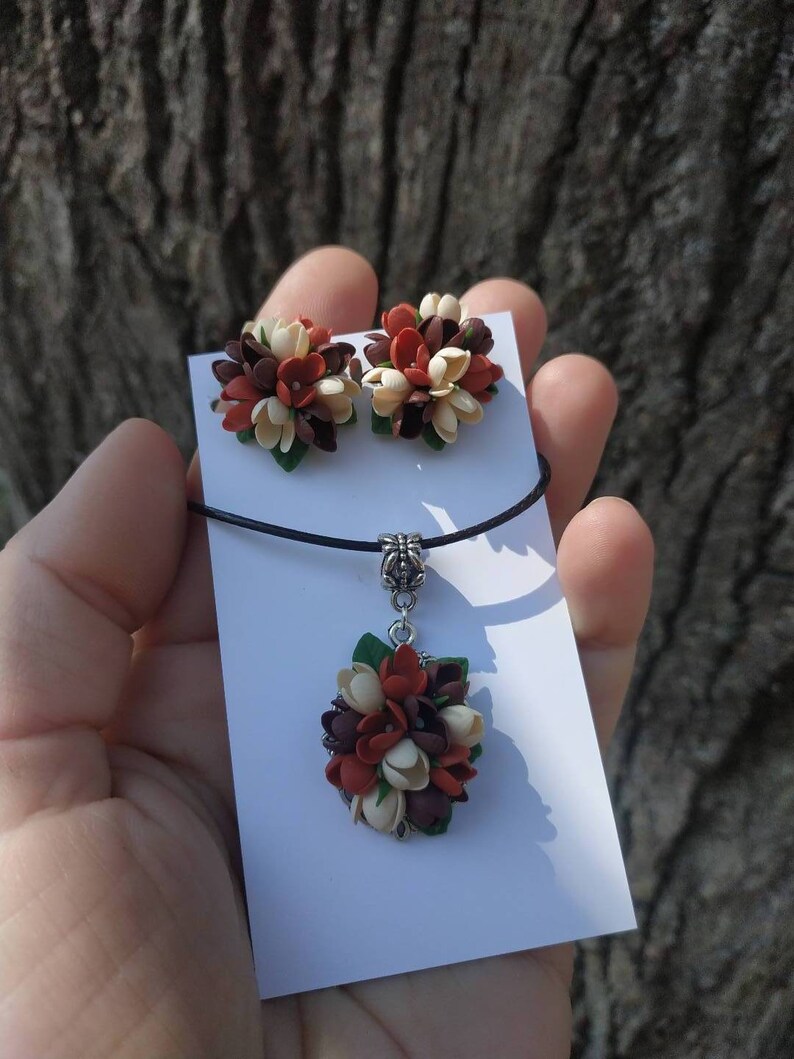May include: A set of handmade jewelry featuring a necklace and earrings. The necklace has a pendant with a cluster of brown, white, and orange flowers. The earrings are also clusters of brown, white, and orange flowers.