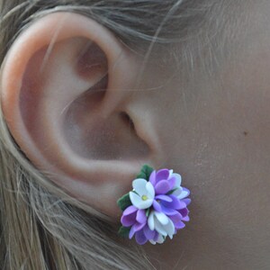 May include: A pair of purple, white, and green flower earrings. The earrings are made of polymer clay and are shaped like a cluster of flowers.
