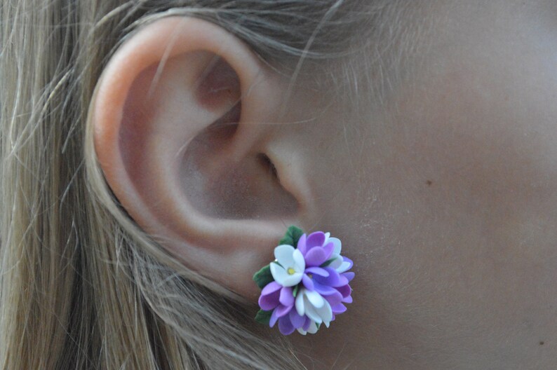 May include: A pair of purple, white, and green flower earrings. The earrings are made of polymer clay and are shaped like a cluster of flowers.