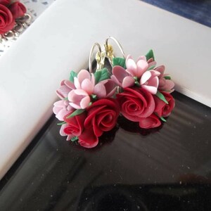 Peut inclure: Une paire de boucles d'oreilles florales compos&eacute;es de grappes de roses rouges sculpt&eacute;es et de fleurs rose p&acirc;le, agr&eacute;ment&eacute;es de petites feuilles vertes. Les boucles sont fix&eacute;es &agrave; des crochets en m&eacute;tal argent&eacute;, r&eacute;v&eacute;lant un design botanique d&eacute;taill&eacute;.