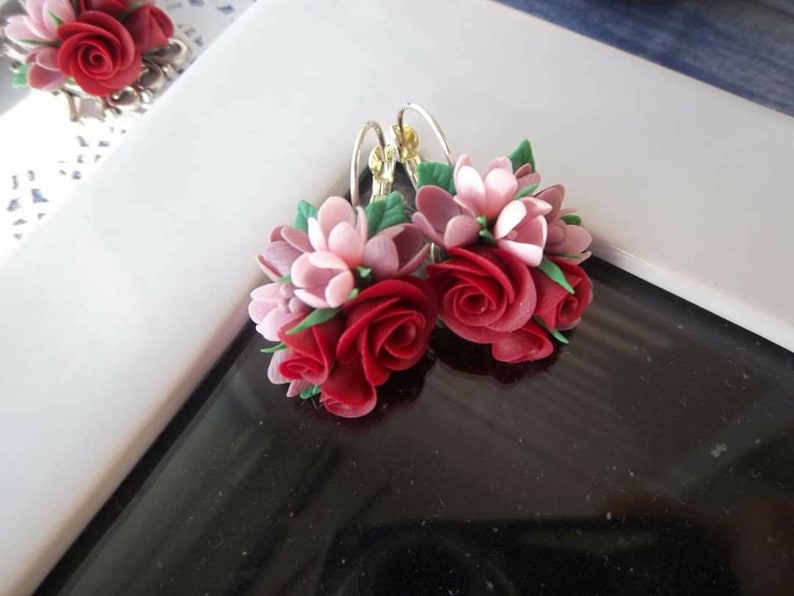 Peut inclure: Une paire de boucles d'oreilles florales compos&eacute;es de grappes de roses rouges sculpt&eacute;es et de fleurs rose p&acirc;le, agr&eacute;ment&eacute;es de petites feuilles vertes. Les boucles sont fix&eacute;es &agrave; des crochets en m&eacute;tal argent&eacute;, r&eacute;v&eacute;lant un design botanique d&eacute;taill&eacute;.