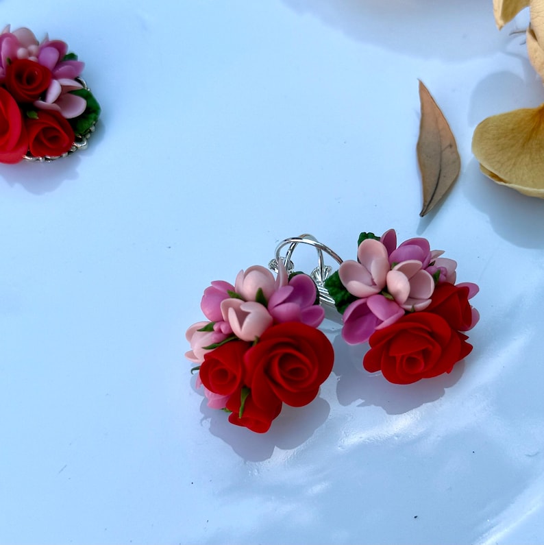 Peut inclure: Une paire de boucles d'oreilles florales compos&eacute;es de grappes de roses rouges et de fleurs rose p&acirc;le avec de petites feuilles vertes. Les boucles d'oreilles sont fix&eacute;es &agrave; des anneaux en m&eacute;tal argent&eacute;. Une troisi&egrave;me pi&egrave;ce assortie est visible en haut &agrave; gauche.