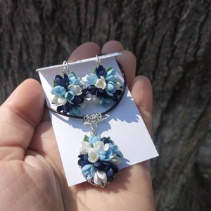 May include: A necklace and earrings set featuring a cluster of blue, white, and dark blue polymer clay flowers. The jewelry is displayed on a white card.