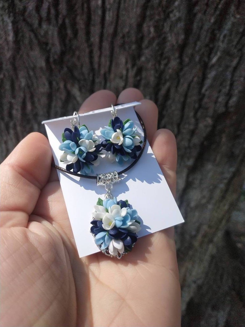 May include: A necklace and earrings set featuring a cluster of blue, white, and dark blue polymer clay flowers. The jewelry is displayed on a white card.