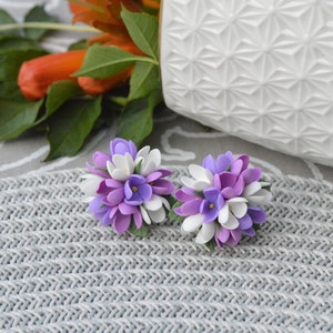 May include: A pair of purple and white flower earrings. The earrings are made of polymer clay and are shaped like small bouquets of flowers.