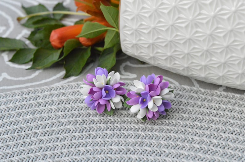May include: A pair of purple and white flower earrings. The earrings are made of polymer clay and are shaped like small bouquets of flowers.