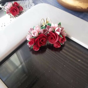 Peut inclure: Une paire de boucles d'oreilles pendantes compos&eacute;es de grappes de roses sculpt&eacute;es rouges et rose clair avec de petites feuilles vertes. Les compositions florales sont fix&eacute;es &agrave; des crochets en m&eacute;tal argent&eacute;, cr&eacute;ant un design botanique d&eacute;taill&eacute;.