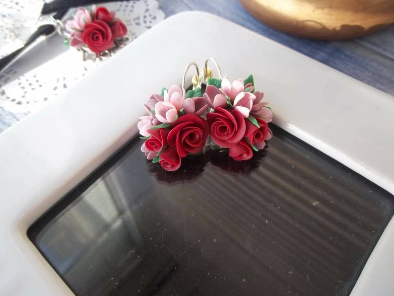 Peut inclure: Une paire de boucles d'oreilles pendantes compos&eacute;es de grappes de roses sculpt&eacute;es rouges et rose clair avec de petites feuilles vertes. Les compositions florales sont fix&eacute;es &agrave; des crochets en m&eacute;tal argent&eacute;, cr&eacute;ant un design botanique d&eacute;taill&eacute;.