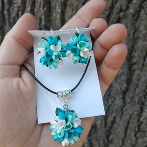 May include: A necklace and earrings set featuring blue and white flower-shaped pendants. The necklace has a black cord and a silver clasp. The earrings have silver hooks.