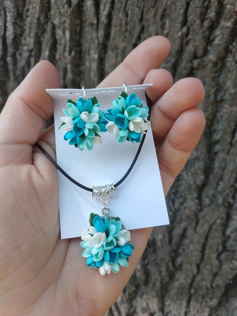 May include: A necklace and earrings set featuring blue and white flower-shaped pendants. The necklace has a black cord and a silver clasp. The earrings have silver hooks.
