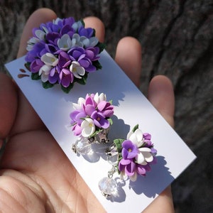 May include: A set of lilac flower earrings with crystal accents. The earrings are made of polymer clay and feature a cluster of purple and white flowers.