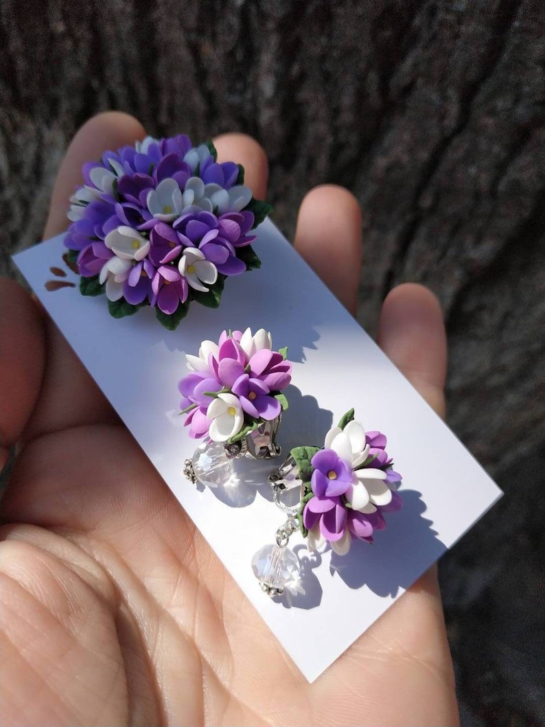 May include: A set of lilac flower earrings with crystal accents. The earrings are made of polymer clay and feature a cluster of purple and white flowers.