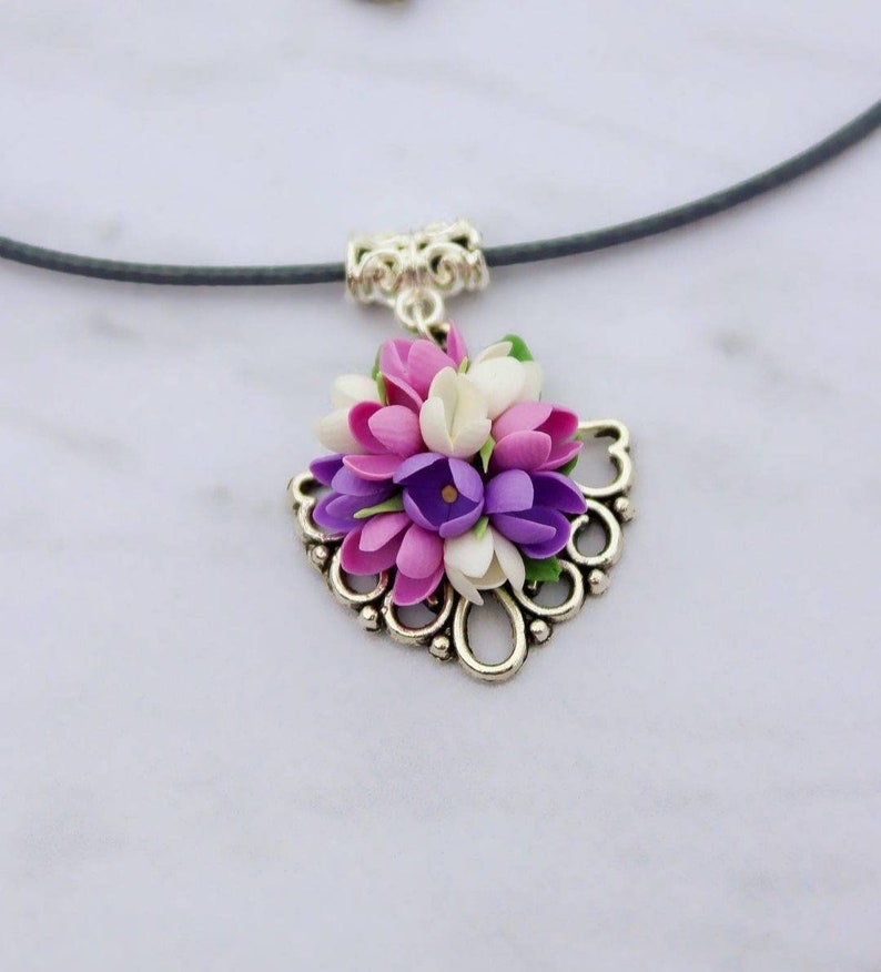 May include: A silver necklace with a floral pendant featuring a cluster of purple, pink, and white flowers. The flowers are arranged in a heart-shaped design and are accented with green leaves.
