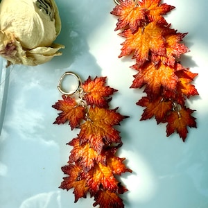 May include: A pair of autumn-themed earrings featuring detailed maple leaf pendants. The leaves display a gradient of colors, from deep red to vibrant orange and yellow, attached to gold-colored hoops. The earrings are designed to dangle.