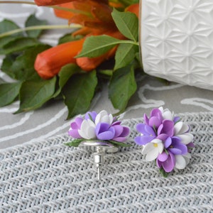 May include: A pair of purple and white flower earrings. The earrings are made of polymer clay and are shaped like small bouquets of flowers. The flowers are a mix of purple and white, with a few yellow centers. The earrings are attached to silver posts.