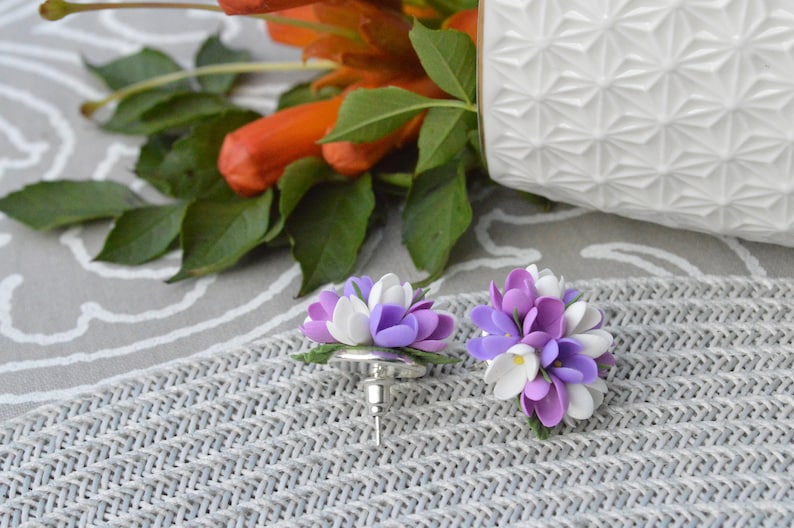 May include: A pair of purple and white flower earrings. The earrings are made of polymer clay and are shaped like small bouquets of flowers. The flowers are a mix of purple and white, with a few yellow centers. The earrings are attached to silver posts.