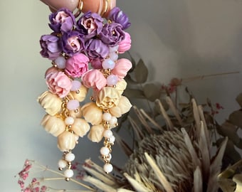 May include: Dangling floral earrings in shades of purple, pink, and cream. The earrings feature clusters of small flower buds, with gold-colored hoops and delicate bead accents. The earrings are held up against a soft background.