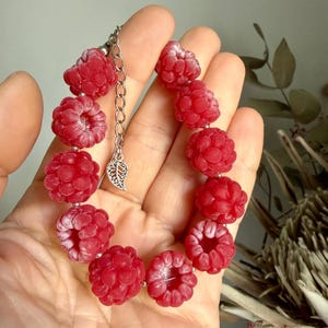 May include: A raspberry-themed bracelet featuring multiple bright red, realistic-looking raspberry beads. The bracelet is held in a hand and has a silver chain and leaf charm. The bracelet is a unique piece of jewelry.