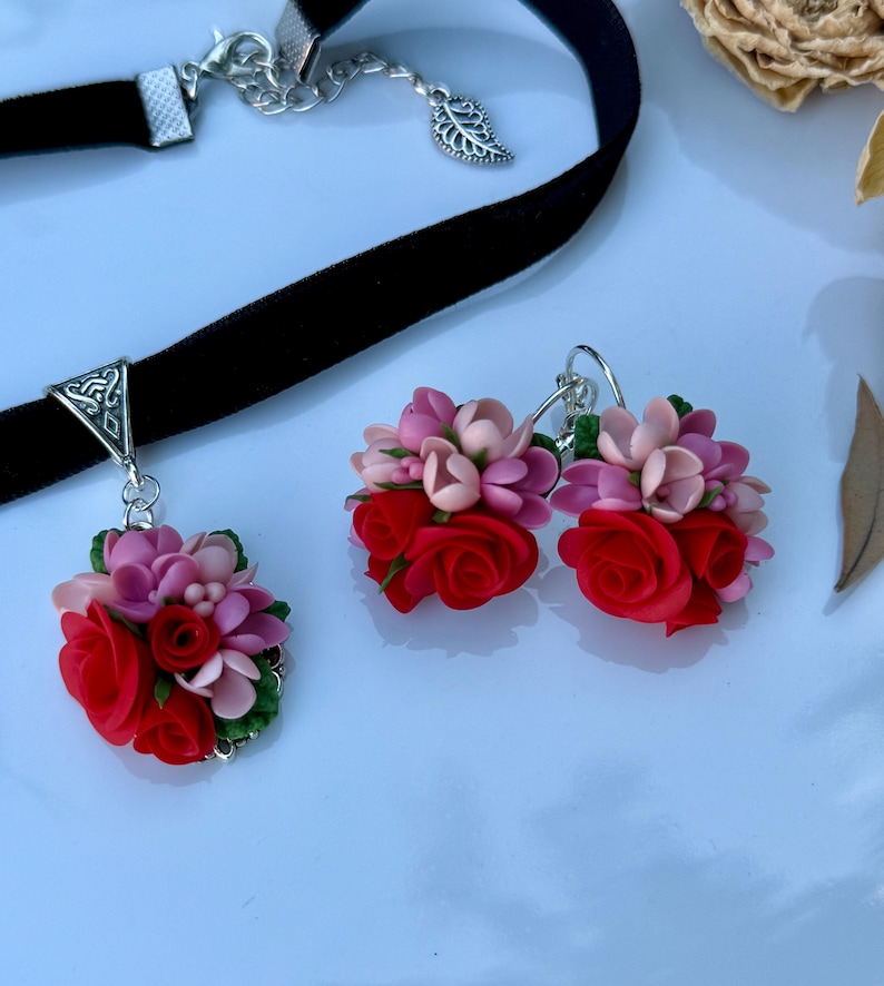 Peut inclure: Un collier ras-du-cou en velours noir avec une b&eacute;li&egrave;re triangulaire argent&eacute;e et un pendentif compos&eacute; de roses rouges et de fleurs roses sculpt&eacute;es avec des feuilles vertes, accompagn&eacute; d'une paire de boucles d'oreilles assorties.
