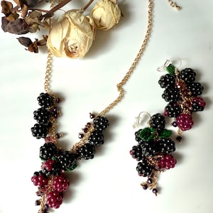 May include: A gold chain necklace and earrings featuring black and burgundy beads, resembling a cluster of blackberries. The necklace has a pendant of blackberries with a cluster of burgundy beads hanging below. The earrings are a matching cluster of blackberries with burgundy beads.