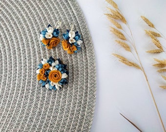 May include: A pair of earrings and a brooch featuring a cluster of white and blue flowers with a yellow center. The jewelry is made of polymer clay.