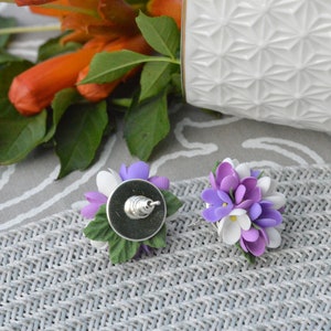 May include: A pair of earrings featuring a cluster of purple and white flowers with green leaves. The earrings are made of polymer clay and have silver posts.