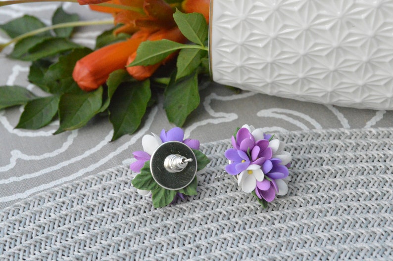 May include: A pair of earrings featuring a cluster of purple and white flowers with green leaves. The earrings are made of polymer clay and have silver posts.