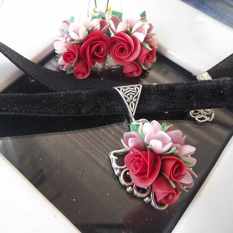 Peut inclure: Un collier ras-du-cou en velours noir et des boucles d'oreilles assorties. Les bijoux pr&eacute;sentent des grappes de roses sculpt&eacute;es rouges et rose clair avec de petites feuilles vertes. Les montures sont en m&eacute;tal argent&eacute; avec un filigrane complexe.