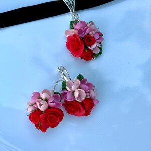 Peut inclure: Un collier ras-du-cou en velours noir avec un pendentif floral et des boucles d'oreilles assorties. Chaque pi&egrave;ce pr&eacute;sente un bouquet de roses sculpt&eacute;es rouges, rose clair et rose fonc&eacute; avec de petites feuilles vertes sur m&eacute;tal argent&eacute;.