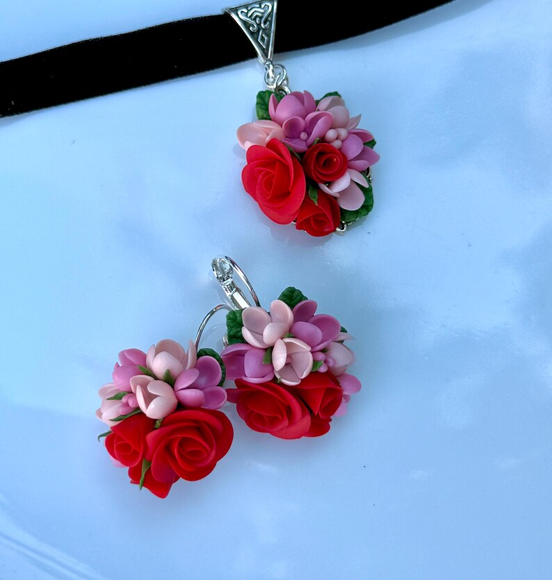 Peut inclure: Un collier ras-du-cou en velours noir avec un pendentif floral et des boucles d'oreilles assorties. Chaque pi&egrave;ce pr&eacute;sente un bouquet de roses sculpt&eacute;es rouges, rose clair et rose fonc&eacute; avec de petites feuilles vertes sur m&eacute;tal argent&eacute;.