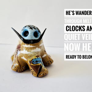 Puede incluir: Una figura de cerámica pintada a mano con un cuerpo marrón claro, ojos azules y un bigote negro. La figura tiene cuernos negros y está decorada con detalles coloridos. Texto: "HE'S WANDERED...READY TO BELONG."