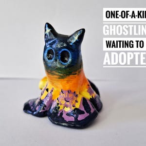 Puede incluir: Figurita de cerámica pintada a mano con forma de gato, con grandes ojos azules. Predominan los colores negro, azul, naranja y morado. El texto dice: "ONE-OF-A-KIND GHOSTLING, WAITING TO BE ADOPTED."