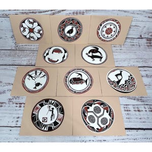 Puede incluir: Diez tarjetas con ilustraciones circulares de estilo tribal en negro, blanco y rojo. Cada tarjeta presenta un diseño único sobre un fondo beige, con motivos geométricos y de animales. Dispuestas sobre una superficie de madera.