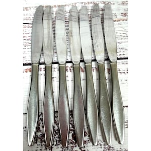 May include: A set of seven silver-toned stainless steel dinner knives arranged in a vertical row. Each knife features a smooth, tapered handle with a matte finish and a polished, slightly curved blade with a serrated edge along one side.