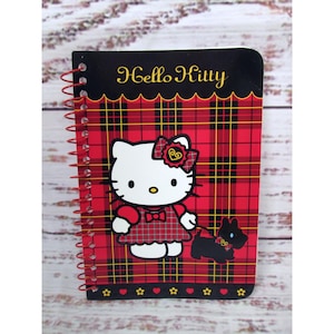 Puede incluir: Un cuaderno de espiral con un patrón de cuadros rojos y negros y un gráfico de Hello Kitty. La portada muestra el texto Hello Kitty, una gata blanca con un vestido y lazo de cuadros, y un pequeño perro negro con correa y detalles en color amarillo.