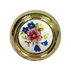 Puede incluir: Un pomo redondo para muebles con un borde metálico de tono dorado y una incrustación central de cerámica blanca. La incrustación está decorada con un patrón floral que consiste en una flor rosa grande, flores azules pequeñas y hojas verdes.