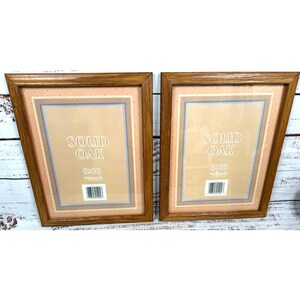 May include: Two rectangular picture frames made of solid oak wood with a natural brown finish. Each frame contains a beige insert with the text SOLID OAK and 8x10 printed in white, along with a barcode label at the bottom center of the insert.