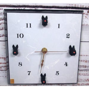 May include: Square wall clock with a white face and black frame. The clock features black Scottie dog accents at the 1, 2, 4, 8, and 11 o'clock positions. The clock hands are gold-colored, and the numbers are black.