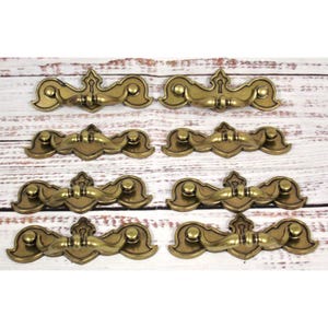 Puede incluir: Ocho tiradores de cajón antiguos de latón con un diseño decorativo. Cada tirador presenta un detalle de ojo de cerradura y un asa curvada. Los tiradores están dispuestos sobre una superficie de madera clara, mostrando su estética vintage.