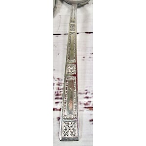 May include: A silver-toned metal spoon handle featuring a geometric pattern with alternating rectangular panels and square sections containing embossed floral motifs. The design is symmetrical, with a smooth, polished finish throughout the length.