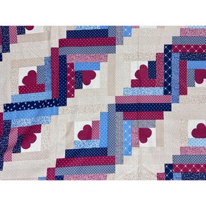 Puede incluir: Un edredón de patchwork con un diseño de cabaña de troncos. El edredón presenta cuadrados con un corazón en el centro. Los cuadrados están rodeados de tiras de tela en tonos de azul, rojo y crema. Las telas tienen varios patrones, incluyendo lunares e estampados florales.