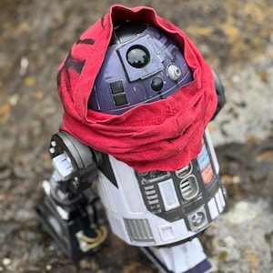 Könnte beinhalten: Eine weiß-violette Roboterfigur mit zylindrischem Körper und kuppelförmigem Kopf. Der Roboter ist in eine rote Stoffkapuze gehüllt, die den oberen Teil des Kopfes bedeckt, wobei die zentrale Linse und die Sensorfelder auf der Kuppel sichtbar bleiben.