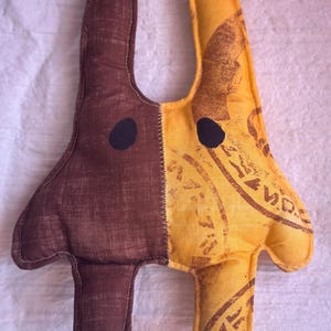 Peut inclure: Une peluche en forme de créature avec deux longues oreilles, présentant un design divisé. Le côté gauche est en tissu marron foncé uni, tandis que le côté droit est en tissu jaune avec des motifs de tampons marron. Elle a deux yeux ovales noirs.