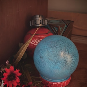May include: A blue decorative sphere with a textured surface sits on a red base. A red sphere with text and a silver top is behind it, along with a geometric container and red flowers.